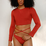 the model is wearing the Doris long-sleeve swim crop top rashguard in terracotta red paired with the Venus striped bikini bottom, a gender-neutral, nonbinary sustainable swimwear set with a soft, modern fit.