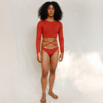 The model is wearing the Doris long-sleeve swim crop top rashguard in terracotta red, paired with the Venus striped terracotta red bikini bottom, it is highlighting the sustainable design and gender-neutral, nonbinary fit.