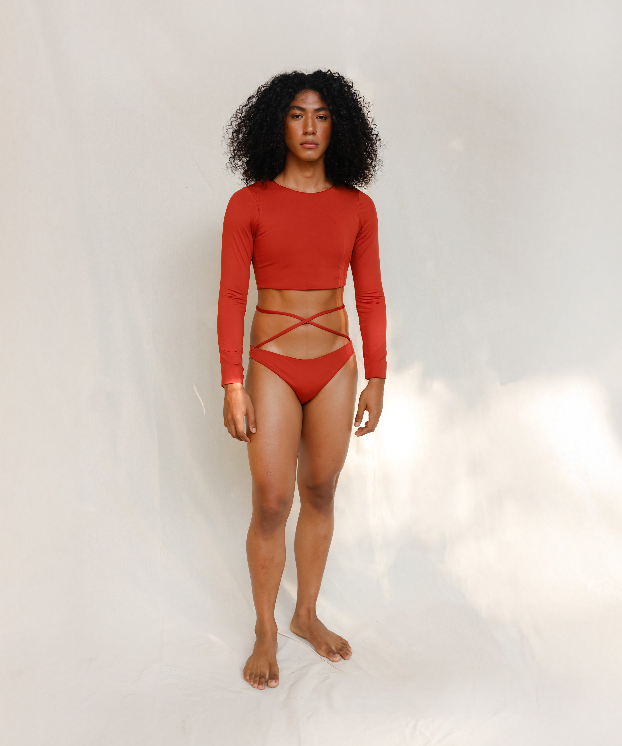 The model is wearing the Doris long-sleeve swim crop top rashguard in terracotta red, paired with the Venus striped terracotta red bikini bottom, it is highlighting the sustainable design and gender-neutral, nonbinary fit.