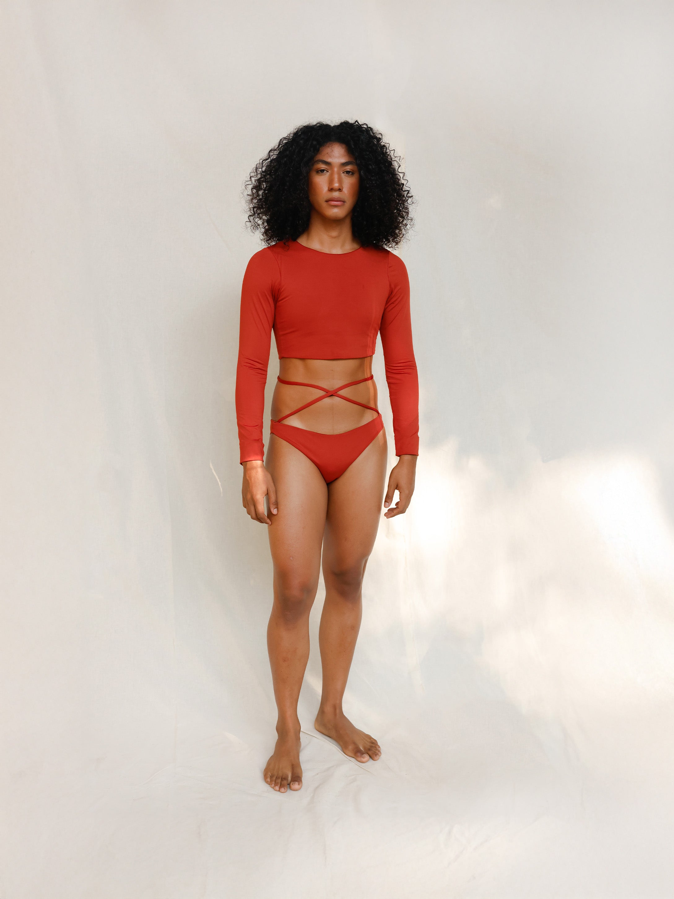 The model is wearing the Doris long-sleeve swim crop top rashguard in terracotta red, paired with the Venus striped terracotta red bikini bottom, it is highlighting the sustainable design and gender-neutral, nonbinary fit.