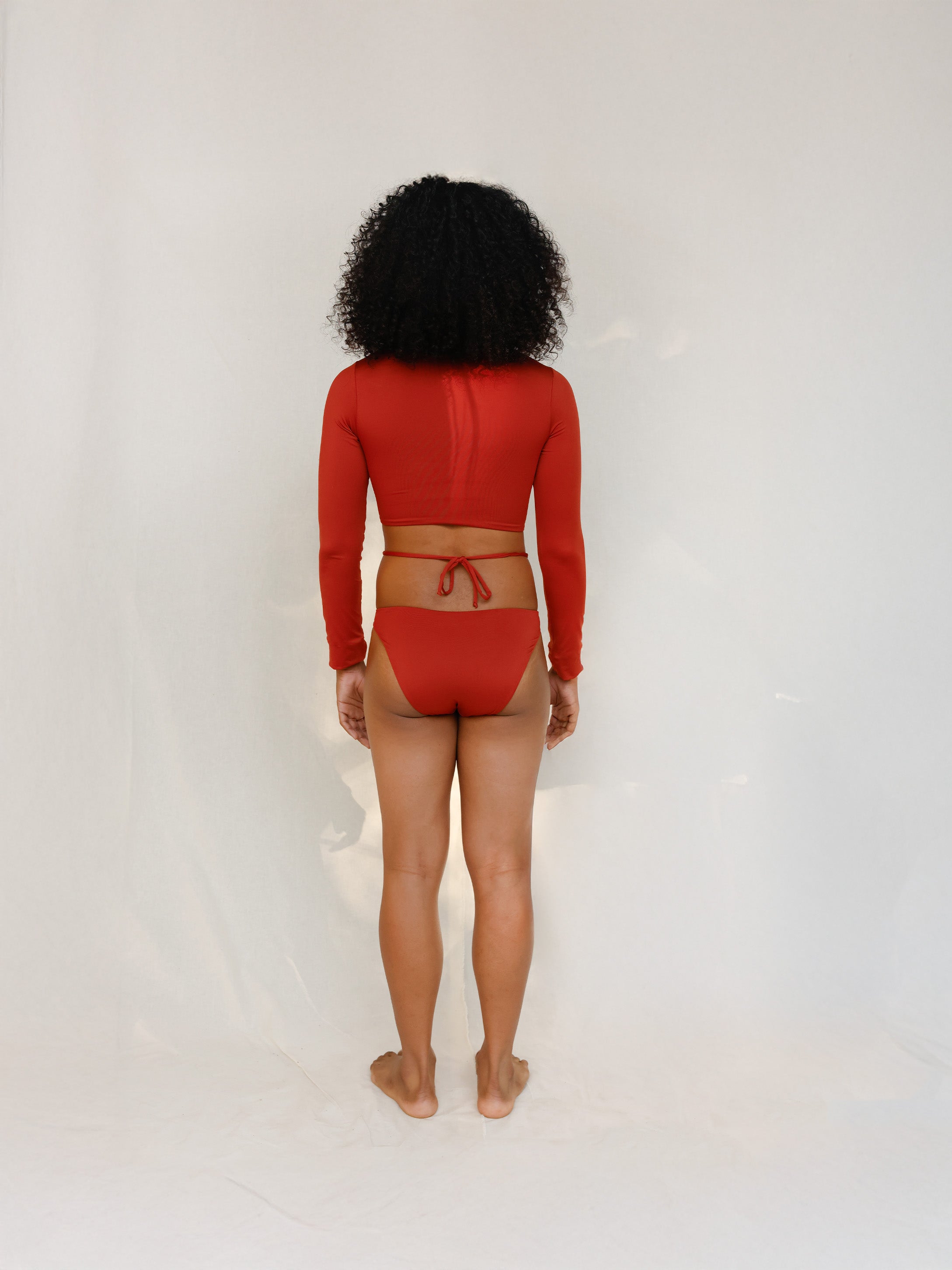 Model wearing non binary terracotta red long-sleeve crop swim top and matching striped bikini bottom designed for secure anatomical coverage and a gender-affirming tuck-friendly fit. 