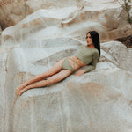 A model is shown from the front wearing the Doris long-sleeve sustainable swim crop top rashguard in mineral green with the Venus striped bikini bottom, a gender-neutral, nonbinary swimwear set with a sleek, modern look.