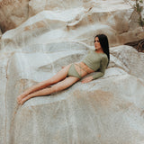 A model is shown from the front wearing the Doris long-sleeve sustainable swim crop top rashguard in mineral green with the Venus striped bikini bottom, a gender-neutral, nonbinary swimwear set with a sleek, modern look.