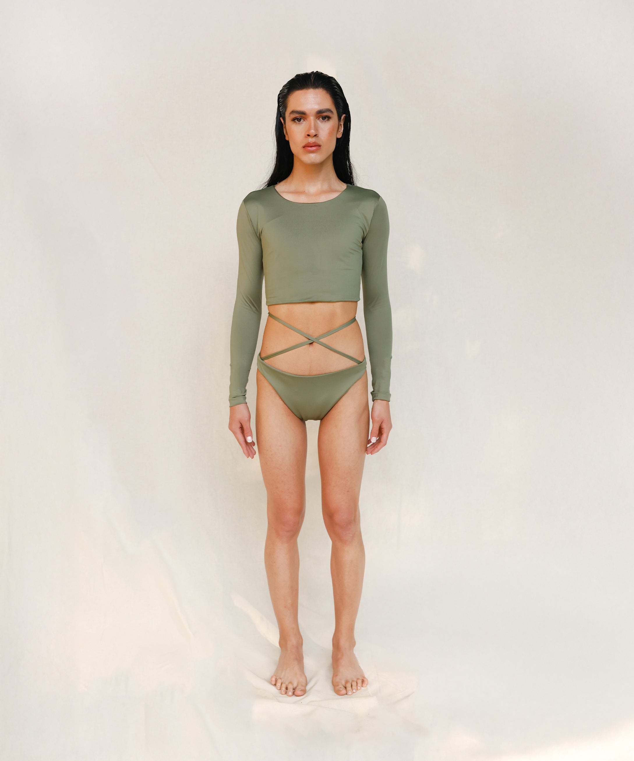 Model wears the Doris long-sleeve swim crop top rashguard in mineral green. It is paired with the Venus striped bikini bottom, a gender-neutral, nonbinary sustainable set shown from the front.