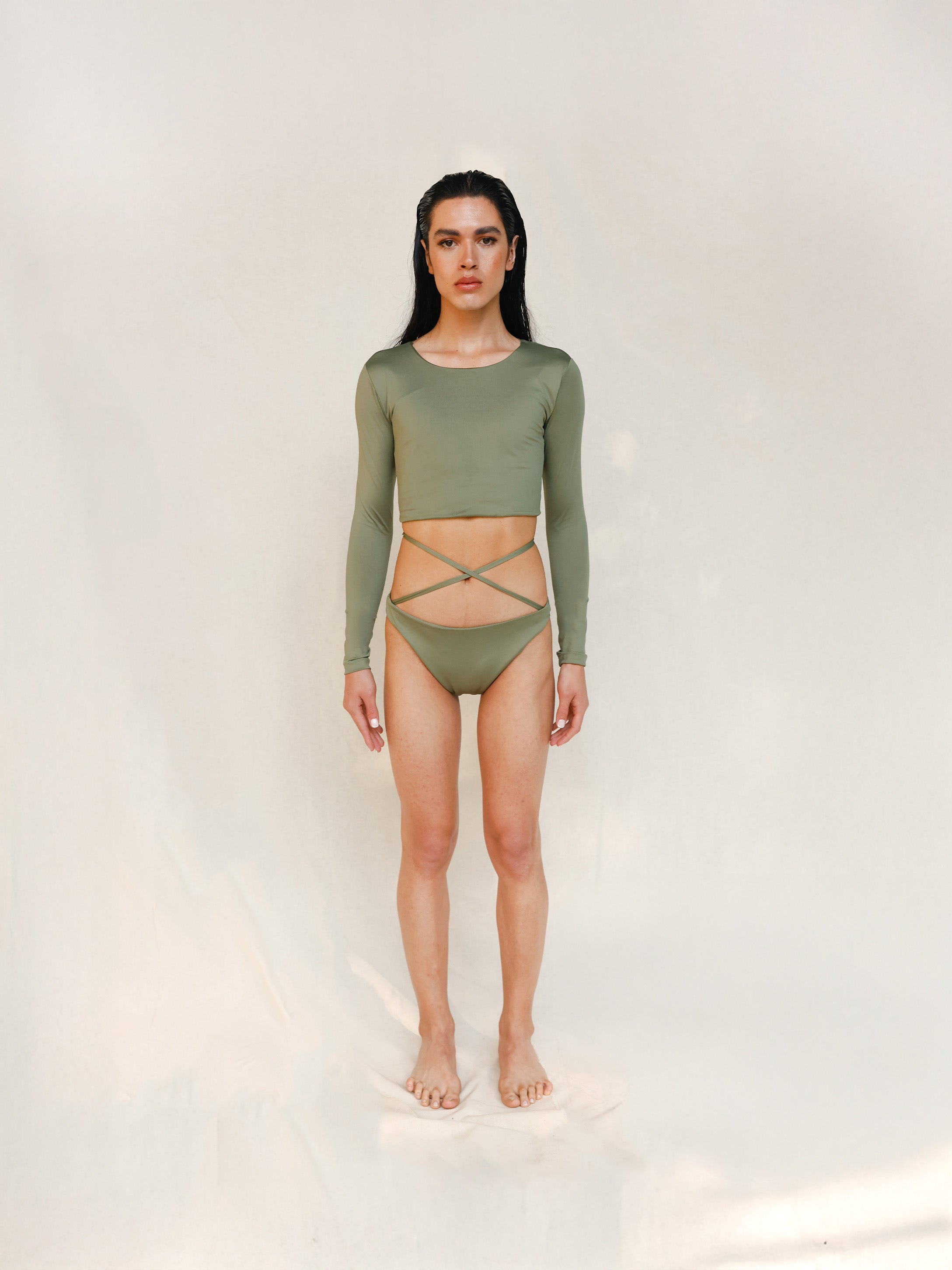 Model wears the Doris long-sleeve swim crop top rashguard in mineral green. It is paired with the Venus striped bikini bottom, a gender-neutral, nonbinary sustainable set shown from the front.