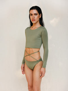 The model is wearing the Doris long-sleeve swim crop top rashguard in mineral green with the Venus striped bikini bottom, shown from the side as a gender-neutral, nonbinary sustainable swimwear set.