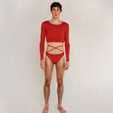 The model wears the Doris long-sleeve swim crop top rashguard in terracotta red with the Venus striped bikini bottom, a gender-neutral, nonbinary swimwear look shown from the front.