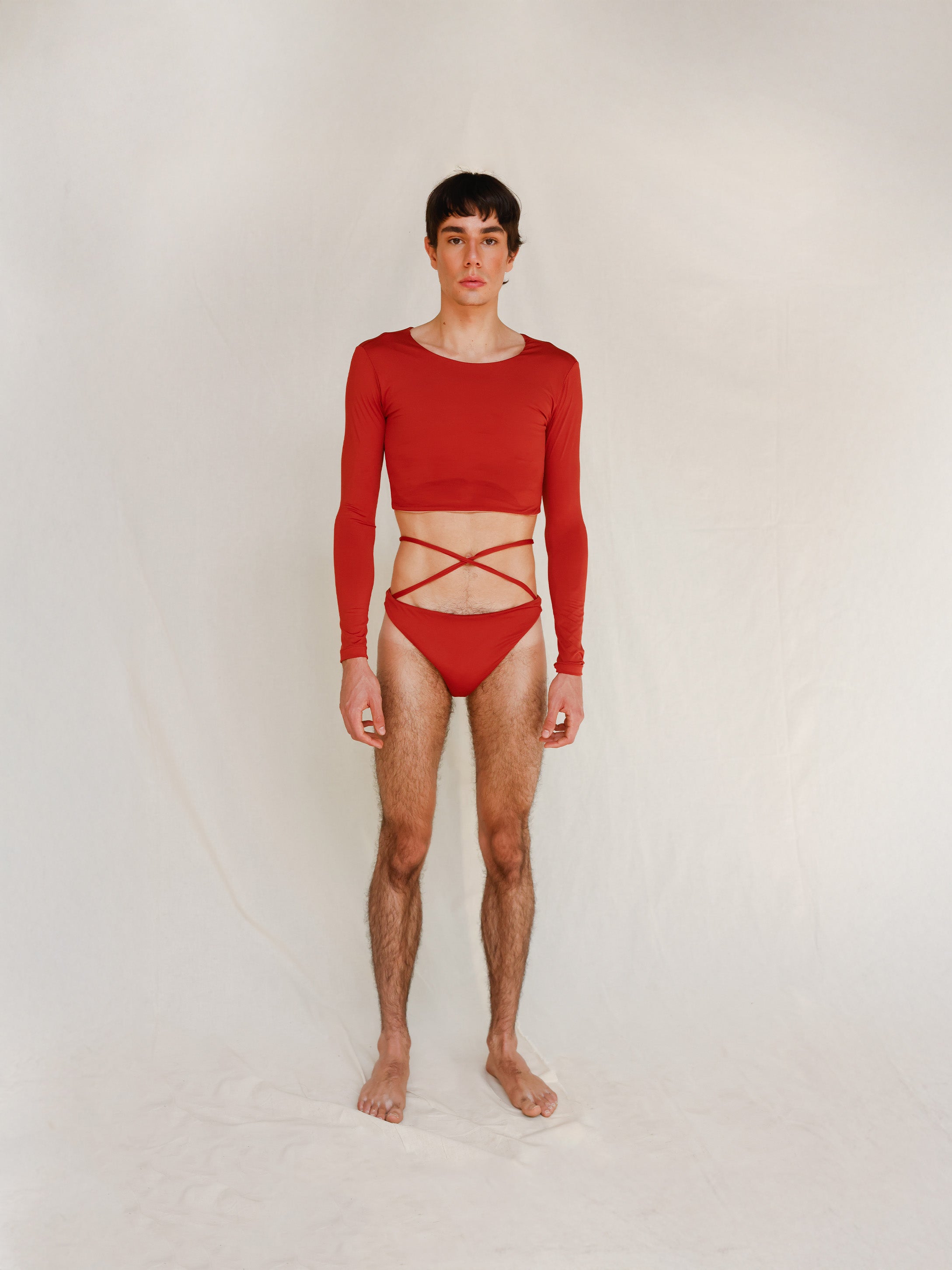 The model wears the Doris long-sleeve swim crop top rashguard in terracotta red with the Venus striped bikini bottom, a gender-neutral, nonbinary swimwear look shown from the front.