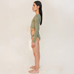 The model is styled in the Doris long-sleeve swim crop top rashguard in mineral green with the Venus striped bikini bottom, shown from the side in a gender-neutral, nonbinary sustainable swimwear look.