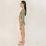 The model is styled in the Doris long-sleeve swim crop top rashguard in mineral green with the Venus striped bikini bottom, shown from the side in a gender-neutral, nonbinary sustainable swimwear look.