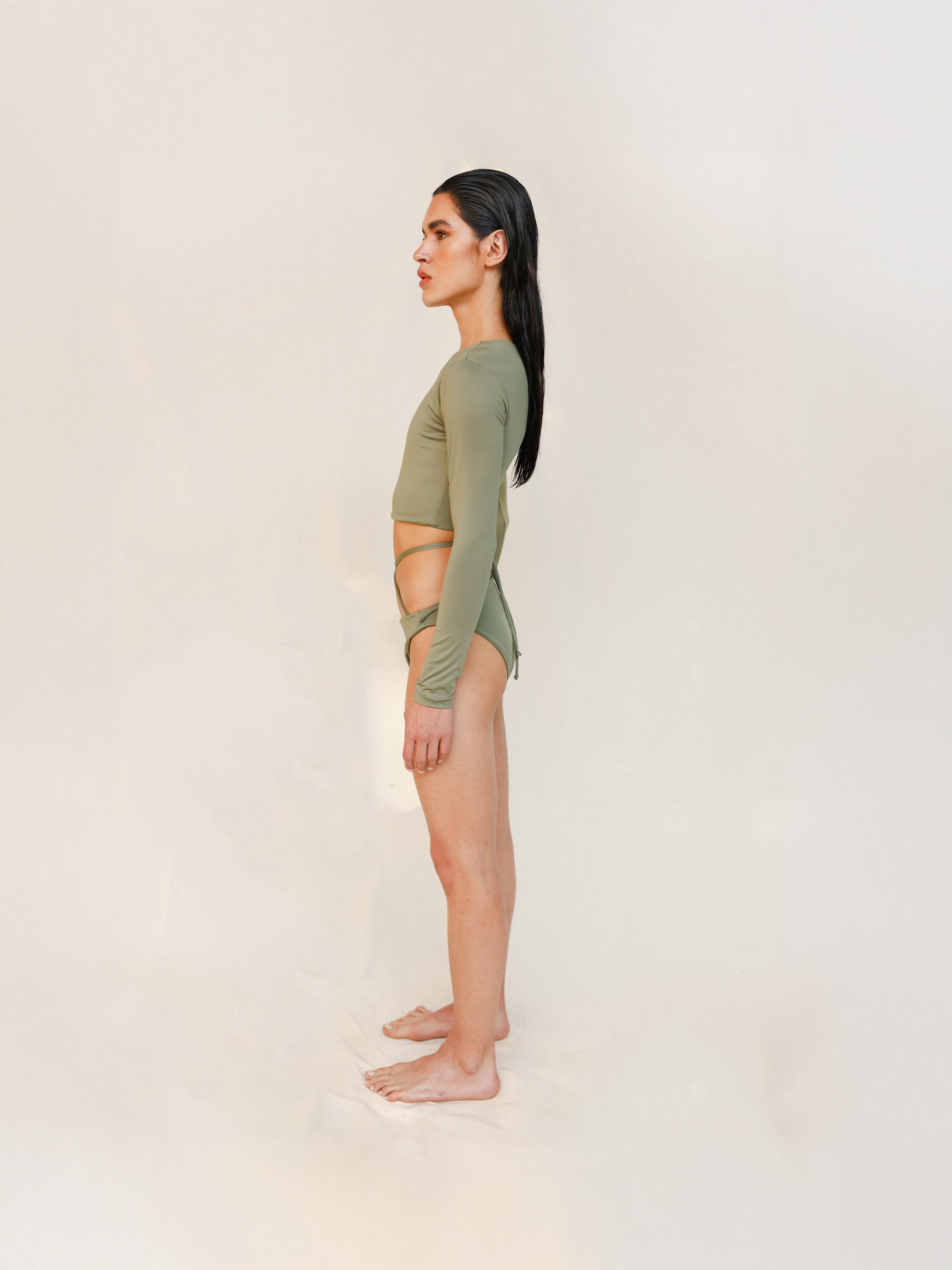 The model is styled in the Doris long-sleeve swim crop top rashguard in mineral green with the Venus striped bikini bottom, shown from the side in a gender-neutral, nonbinary sustainable swimwear look.